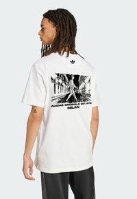 Biele bavlnené tričko s čiernym grafickým potlačou na chrbte, znázorňujúcim uličnú scénu s osobou, ktorá kráča. Logo Adidas na hornej časti chrbta.