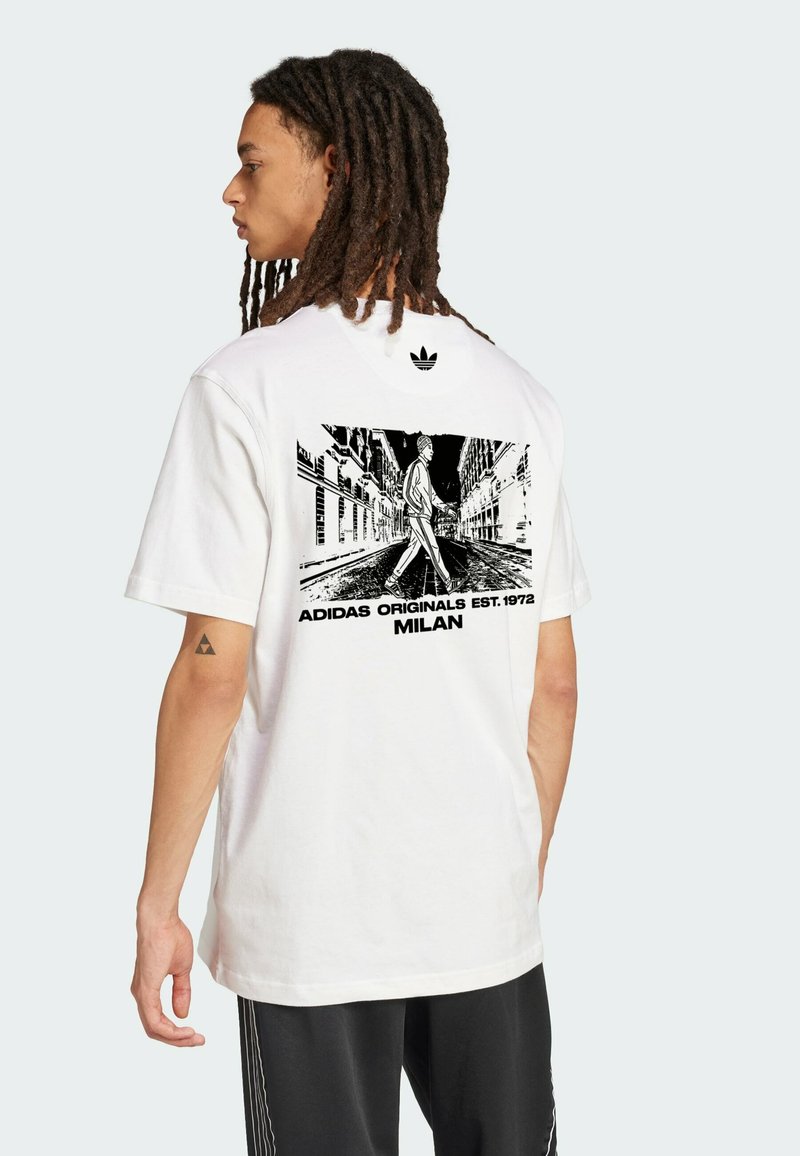 Bijela pamučna majica s crnim grafičkim printom na leđima, koja prikazuje ulično okruženje s osobom koja hoda. Adidas logo na gornjem dijelu leđa.