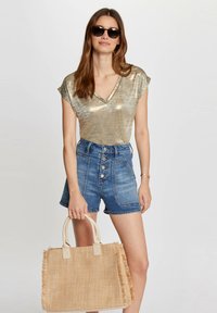 Metallisk guld V-hals top, højhvide denim shorts med frontknapper og en tan stråtask med frynser. Solbriller inkluderet.