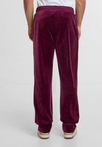 Mann mit tief lila Samthose und cremefarbenen Sneakern, von hinten vor einem schlichten weißen Hintergrund gezeigt.