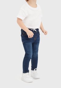 Blå denimjeans med slim fit, elastisk midja, fem fickor och kontrasterande sömmar, kombinerat med en enfärgad vit t-shirt med kort ärm och vita sneakers.