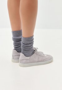Sneakers din suede gri deschis cu talpă albă, asortați cu șosete gri cu model în dungi, creând un contrast de textură stratificat împotriva pielii goale.