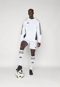 Ensemble de sport Adidas comprenant un sweat à capuche blanc avec des accents noirs, un short blanc et des chaussettes rayées, assortis à des crampons noirs et un ballon de football multicolore.
