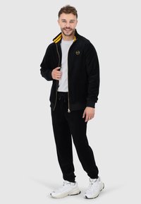 Schwarzer Velours-Trainingsanzug mit goldenen Akzenten, bestehend aus einer Reißverschlussjacke mit gestreiftem Kragen, elastischen Bundhosen und weißen Sportschuhen.