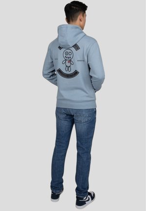 Sudadera azul clara con un gran gráfico negro que presenta una muñeca y texto en la parte de atrás, combinada con jeans azul oscuro y zapatillas negras.