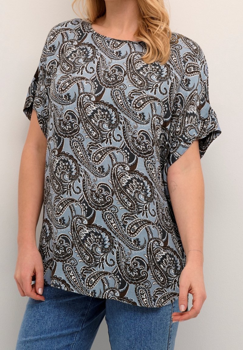Lys blå bluse med et paisley mønster i sort og hvid. Korte ærmer og en løs pasform, lavet af et blødt, tekstureret stof.