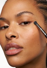 Pennello per il trucco che applica il prodotto vicino all'occhio, vista ravvicinata di una pelle liscia, colore naturale delle labbra e sopracciglia definite. Sfondo neutro.