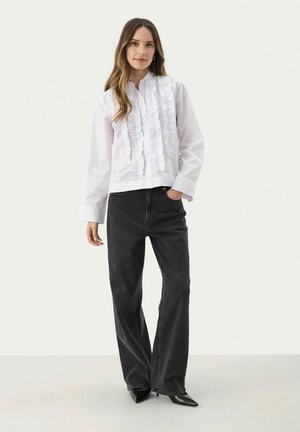 White cotton blouse with ruffled front detail, paired with black straight-leg jeans. Model wears pointy black heels.