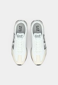 Fekete és szürke díszítéssel ellátott fehér tornacipők. Szintetikus anyagból készültek, fűzőkkel, lekerekített orral és texturált gumitalppal.