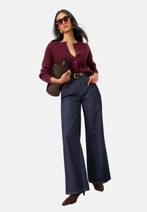 Frau trägt übergroßen bordeauxroten Strickcardigan, hoch taillierte, weit geschnittene dunkle Jeans, braune spitze Stiefel, große Sonnenbrille und hält eine braune Clutch.