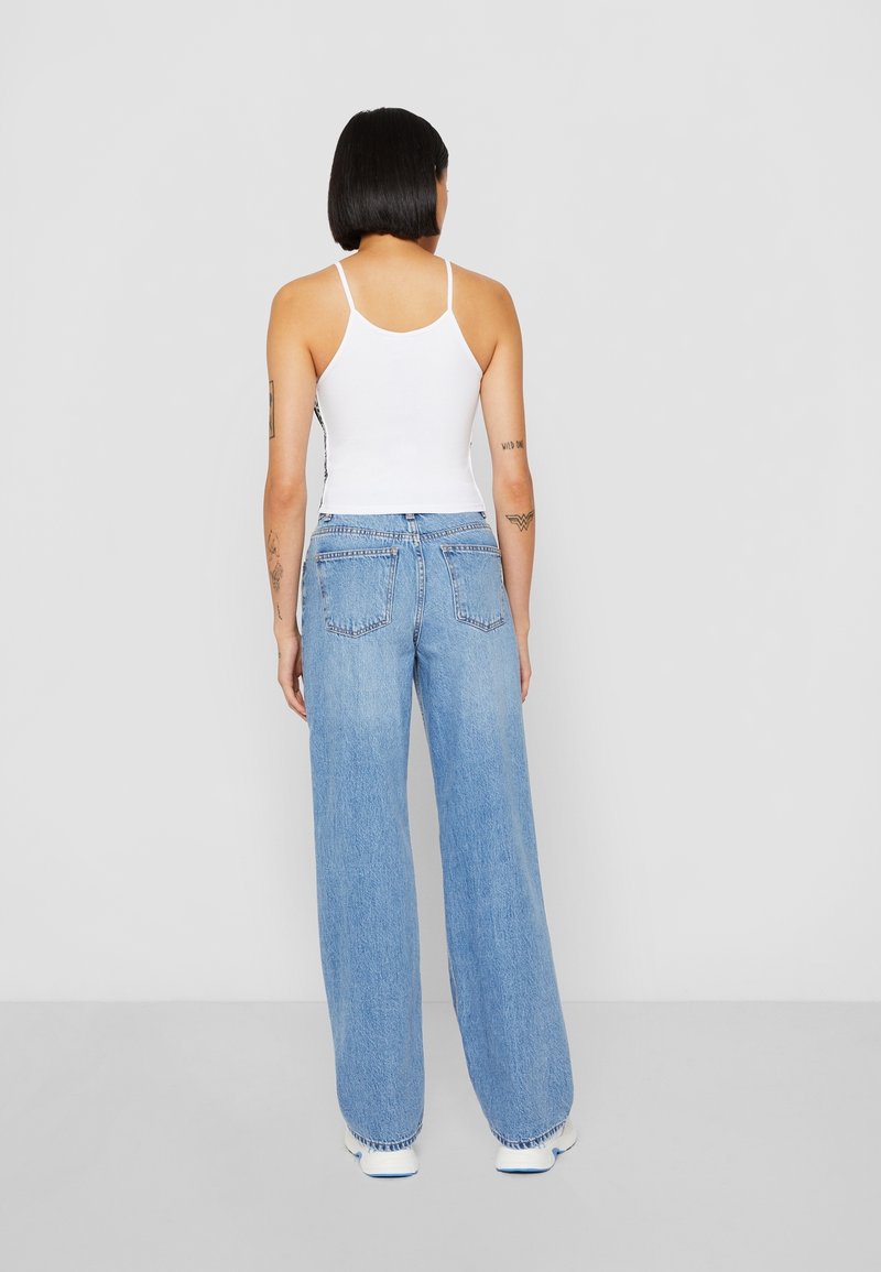Femme aux cheveux noirs courts portant un débardeur blanc, un jean large bleu clair et des baskets blanches, debout face à un fond uni gris clair.
