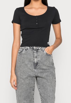 Musta lyhythihainen toppi, jossa on pyöreä kaula-aukko ja kolmesta napista koostuva nappilista, yhdistettynä korkeavyötäröisiin harmaisiin farkkuihin.