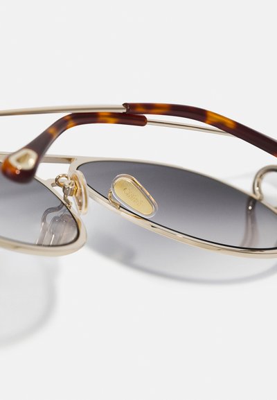 Óculos de sol de metal com lentes cinzas, apresentando uma armação dourada e braçadeiras em padrão tartaruga. Detalhe notável inclui um logótipo na lente.