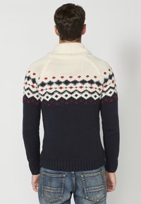 Suéter de punto con un patrón fair isle en crema, azul marino y rojo en la parte superior, con un cuello alto y un dobladillo acanalado.
