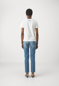 Biała koszulka z krótkim rękawem z logo "MOSCHINO" na plecach, zestawiona z jasnoniebieskimi jeansami o prostych nogawkach i czarnymi klapkami na obcasie.