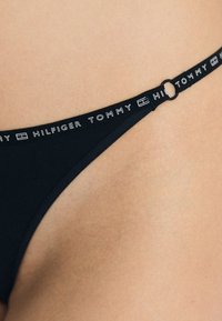 Musthave mustra mustra mustra, selline musta tooni stringkätisega, millel on logo riba "TOMMY HILFIGER", ümmargune detail ja sile tekstuur. Minimalistlik disain, mugav istuvus.