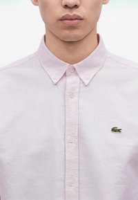 Homme portant une chemise à boutons rose clair avec un petit logo crocodile vert sur la poitrine gauche.