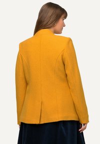 Giacca in misto lana gialla con vestibilità su misura, dotata di colletto alto, spacco posteriore e dettagli cuciti. Tessuto testurizzato, design minimalista.