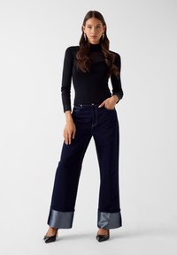 Frau trägt ein schwarzes, eng anliegendes Langarmshirt, weite dunkle Jeans mit großen umgeschlagenen Bündchen, schwarze spitze Absätze und baumelnde Ohrringe und steht.