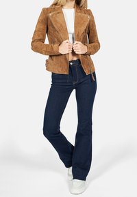 Bruine suède motorjas met ritsdetails, hoge kraag en zijgespen, gecombineerd met donkerblauwe uitlopende jeans en witte sneakers.