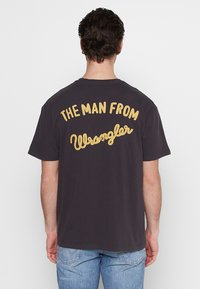 Camiseta de algodón de color oscuro con un gran texto amarillo bordado en la parte de atrás que dice "EL HOMBRE DE Wrangler." Corte casual.