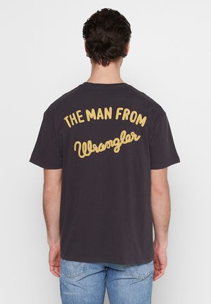 Camiseta de algodón de color oscuro con un gran texto amarillo bordado en la parte de atrás que dice "EL HOMBRE DE Wrangler." Corte casual.