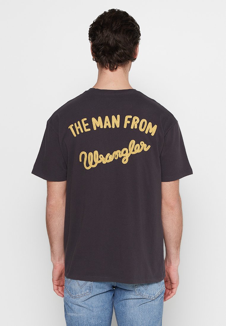 Camiseta de algodón de color oscuro con un gran texto amarillo bordado en la parte de atrás que dice "EL HOMBRE DE Wrangler." Corte casual.
