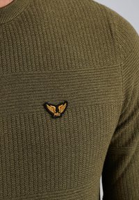 Olivovo zelený pletený sveter s textúrovanými vertikálnymi a horizontálnymi pruhmi. Na hrudi má čiernu nášivku so zlatým krídlovým emblémom.
