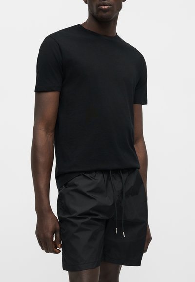 Άνδρας που φοράει απλό μαύρο κοντομάνικο T-shirt και μαύρα σορτς με κορδόνι σε ουδέτερο φόντο.