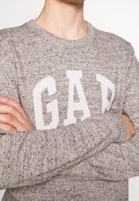 Das graue Strickpullover verfügt über einen runden Ausschnitt und das auffällige weiße "GAP"-Logo. Der Stoff hat eine strukturierte Oberfläche in unterschiedlichen Grautönen.