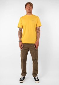 T-shirt en coton jaune avec un col rond et des manches courtes, associé à un pantalon cargo olive. Des baskets noir et blanc complètent la tenue.