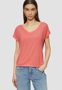 Korallenfarbendes V-Ausschnitt-T-Shirt mit kurzen Ärmeln aus leichtem Stoff. Es hat eine lockere Passform mit aufgerollten Ärmeln und einer glatten Textur. Kombiniert mit hellblauen Jeans.