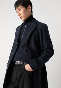 Sobretudo de lã azul-marinho com lapelas picadas, sobre um pulôver azul-marinho com zip e jeans pretos, apresentando um cinto texturizado e uma silhueta elegante.