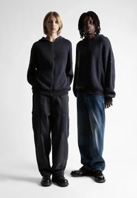 Două persoane stau umăr la umăr, purtând hanorace negre cu glugă și fermoar, pantaloni largi - unul în negru, celălalt în blugi - și pantofi negri.