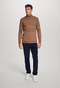 Bruine gebreide coltrui met gestructureerde banden, gecombineerd met marineblauwe broek en witte sneakers. Casual outfit met een aansluitende silhouet.