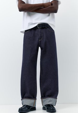 Osoba stojí so založenými rukami, oblečené tmavé široké džíny s vyhrnutými nohavicami, čierny opasok, čierne kožené topánky a biela košeľa s krátkym rukávom.