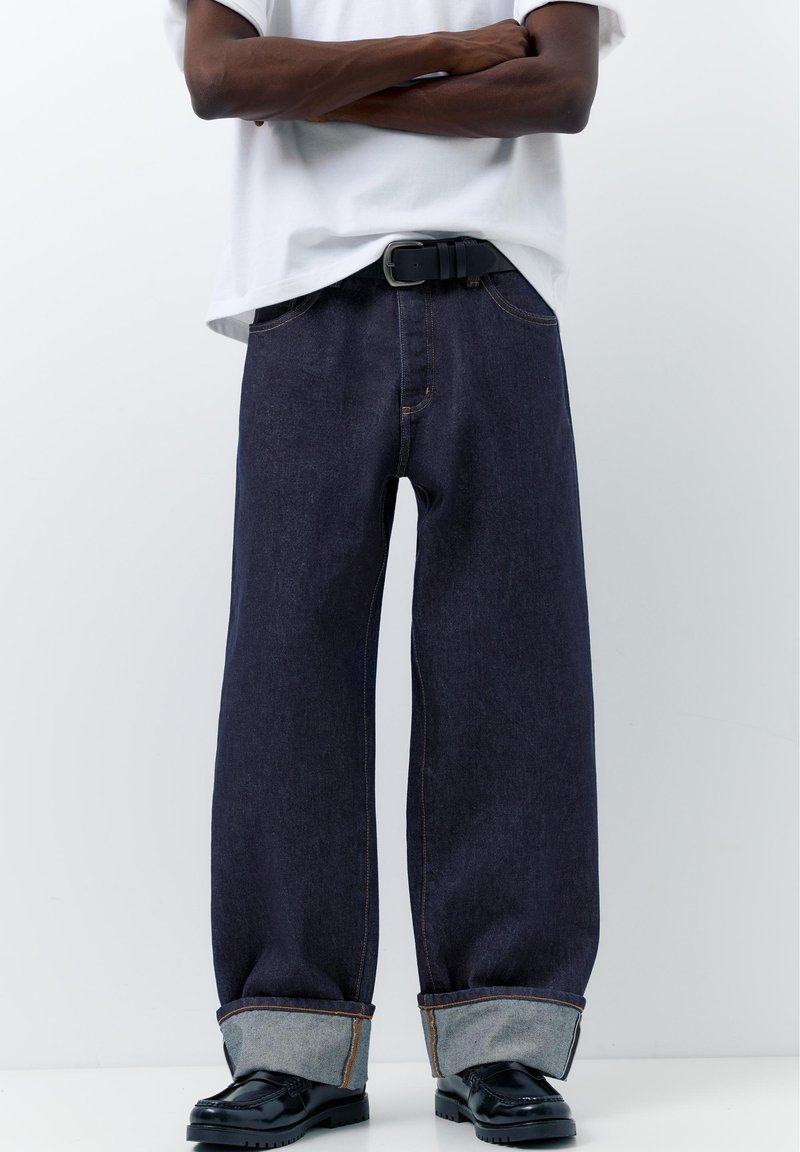 Personne debout, les bras croisés, portant un jean large foncé avec revers, une ceinture noire, des chaussures en cuir noires et une chemise blanche à manches courtes.