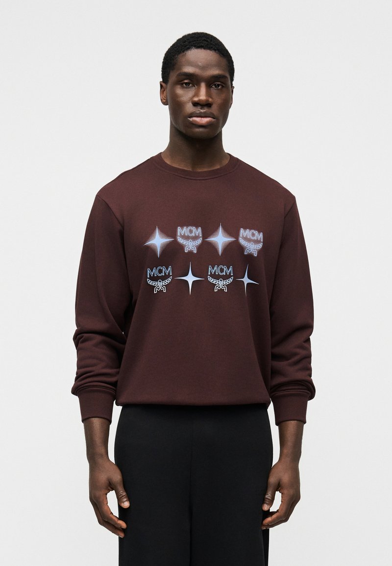 Καφέ βαμβακερό φούτερ με μεγάλα λευκά και μπλε λογότυπα MCM και μοτίβα αστεριών στο μπροστινό μέρος. Ριμπ μανσέτες και λαιμόκοψη, άνετη εφαρμογή.