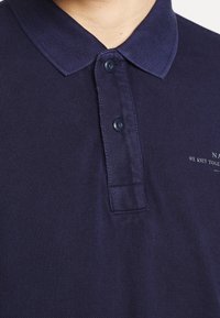Marineblå polo-shirt lavet af tekstureret stof med klassisk krave, to-knaps placket og et lille broderet logo på brystet.
