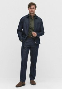 Man som bär en mörkblå jacka och byxor med en olivfärgad skjorta, står med ena handen i fickan och den andra justerar jackans dragkedja.