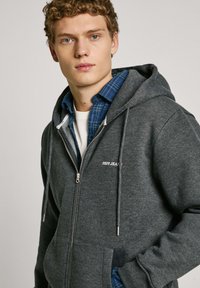Sudadera gris con cremallera, hecha de material suave, con un bolsillo frontal, capucha con cordón y el logo de "Pepe Jeans" en el lado izquierdo del pecho.