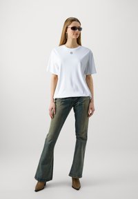 White short-sleeve t-shirt with a small black logo, paired with faded blue flared jeans and brown pointed-toe boots. Sunglasses and hoop earrings.