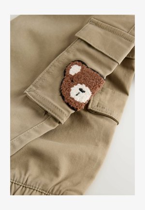 Khaki-kangaskangasta, jossa on tekstuuripintainen ruskea karhu-ompelus; karhu sisältää valkoisia yksityiskohtia ja se on ommeltu vaatteen hihansuuhun.