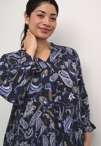Blusa de color azul marino con estampado de motivos paisley y florales. Presenta un escote en V, mangas largas con puños fruncidos y un corte holgado.