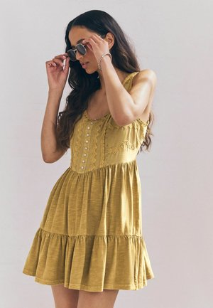 Kurzes gelbes Kleid mit einem gestuften Rock, verziert mit Spitze, Knöpfen auf der Vorderseite und einer weichen Textur. Sonnenbrille in der Hand gehalten.