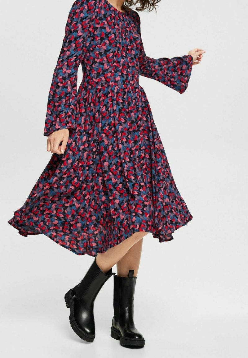 Femme portant une robe à manches longues, longueur genou, à motifs rouges, roses et bleus avec des manches évasées, associée à des bottines noires épaisses mi-mollet.
