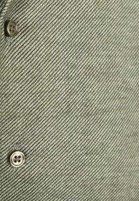 Knopenblouse gemaakt van gestructureerde, lichtgroene stof met een schuin streeppatroon. Beschikt over een bruine knoopdetail bij de kraag.