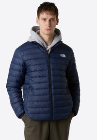 Giacca puffer blu navy con design trapuntato, zip frontale e tasche laterali. Presenta uno strato con cappuccio grigio sotto. Leggera e isolante.