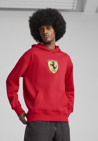 Rød hættebluse med kengurulomme, der har et gult Ferrari-logo. Glat stof, lange ærmer og ribbede manchetter.