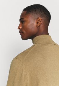 Strikket sweater i beige med høj krave. Har ribbet tekstur og sømdetaljer. Vist i en sideprofil med en glat stoffinish.